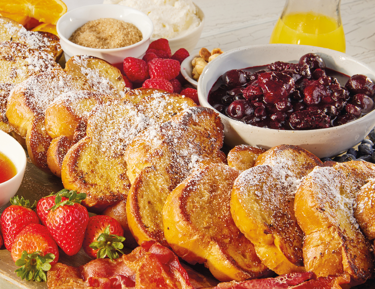 Ten pieces of French Toast with powdered sugar sprinkled on top, next to bacon, strawberries, raspberries, and berry compote